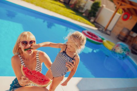 Téléchargez les photos : Belle mère et fille s'amusent à passer une chaude journée ensoleillée en plein air, à manger une tranche de pastèque près de la piscine - en image libre de droit