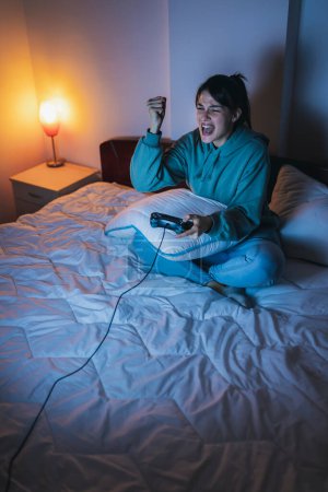 Téléchargez les photos : Belle jeune femme en pyjama s'amusant à la maison, jouant à des jeux vidéo au lit la nuit, excitée après avoir gagné le jeu - en image libre de droit