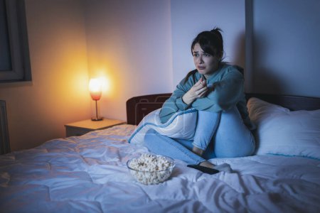 Téléchargez les photos : Vue grand angle de belle jeune femme portant un pyjama assis dans le lit la nuit, triste tout en regardant un film dramatique à la télévision, étreignant l'oreiller et pleurant - en image libre de droit