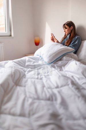 Hermosa joven con pijamas escribiendo mensaje de texto usando un teléfono inteligente en la cama después de despertarse por la mañana