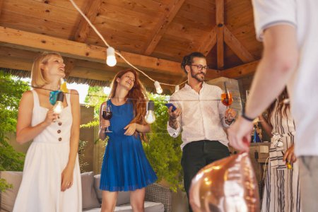Foto de Grupo de jóvenes alegres teniendo una fiesta de cumpleaños sorpresa para un amigo, divirtiéndose bailando y bebiendo cócteles coloridos - Imagen libre de derechos