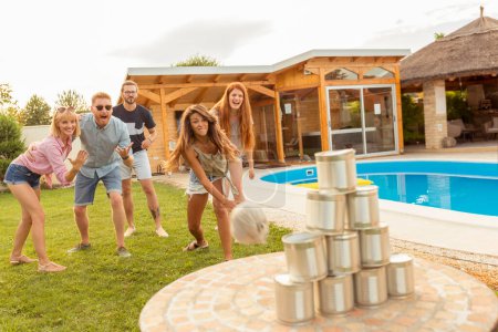 Foto de Grupo de alegres jóvenes amigos que se divierten jugando a las latas derribadas lanzando un juego de pelota mientras están en la fiesta al aire libre junto a la piscina - Imagen libre de derechos
