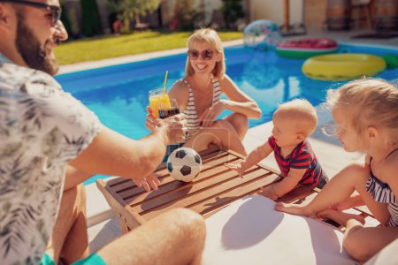 Téléchargez les photos : Belle jeune famille passant une chaude journée ensoleillée au bord de la piscine, parents portant un toast et buvant des cocktails pendant que les enfants jouent - en image libre de droit