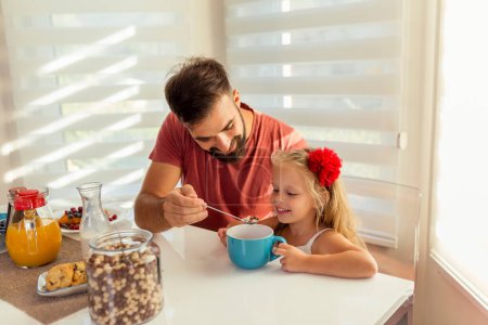 Photo for Young father and beautiful little daughter sitting at dinning room table, having breakfast at home and enjoying their time together - Royalty Free Image
