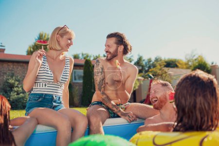 Grupo de jóvenes amigos divirtiéndose en la fiesta de verano en la piscina, sentados junto a la piscina y comiendo paletas de sandía