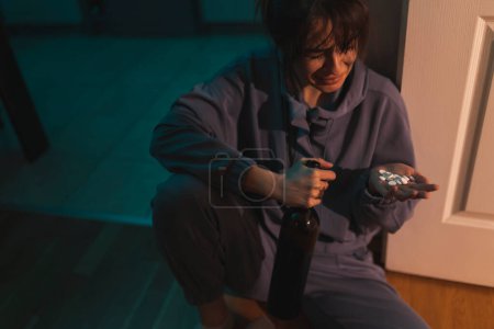 Téléchargez les photos : Une jeune femme désespérée assise sur le sol de la cuisine dans le noir ivre, tenant une bouteille de vin et une poignée de pilules ; abus de substances, dépression et concept de suicide - en image libre de droit