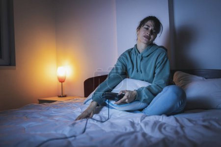 Téléchargez les photos : Belle jeune femme en pyjama s'amusant à la maison, jouant à des jeux vidéo au lit la nuit, déçue après avoir perdu le match - en image libre de droit