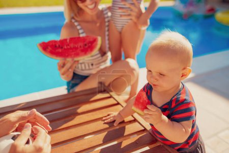 Foto de Los padres jóvenes se divierten pasando el día de verano soleado caliente al aire libre con sus hijos, comiendo rebanadas de sandía junto a la piscina - Imagen libre de derechos