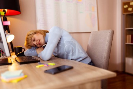 Téléchargez les photos : Femme assise à son bureau à la maison, fatiguée en faisant des heures supplémentaires, s'appuyant sur son bureau et dormant - en image libre de droit