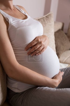 Téléchargez les photos : Gros plan du ventre d'une femme enceinte alors qu'elle est assise sur un canapé dans un salon, et touchant doucement et tenant son ventre - en image libre de droit