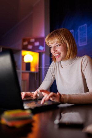 Photo for Woman sitting at her desk in home office working late at night using laptop computer; female web developer working overtime remote from home - Royalty Free Image