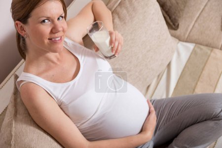 Foto de Hermosa mujer embarazada sentada en un sofá en una sala de estar con un vaso de leche - Imagen libre de derechos