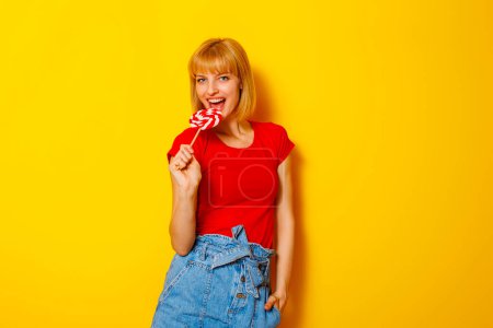 Téléchargez les photos : Belle femme blonde en tenue d'été tendance tenant des bonbons comme micro chantant et dansant sur fond de couleur jaune - en image libre de droit