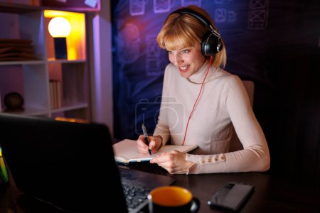 Photo for Woman sitting at her desk in an office working overtime, wearing headset while having conference call and taking notes in planner - Royalty Free Image