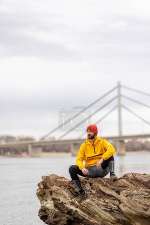 Téléchargez les photos : Homme assis sur un tronc d'arbre tombé, faisant une pause en faisant du jogging au bord de la rivière un jour d'automne couvert, regardant la distance péniblement - en image libre de droit