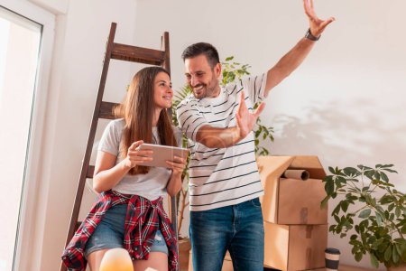 Téléchargez les photos : Couple en amour avoir du plaisir tout en déménageant dans une nouvelle maison, à la recherche d'idées de redécoration appartement à l'aide de tablette - en image libre de droit