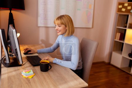 Téléchargez les photos : Femme travaillant à distance de la maison assis à son bureau à la maison en utilisant un ordinateur de bureau - en image libre de droit