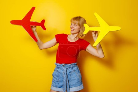 Foto de Hermosa mujer rubia alegre en ropa de verano de moda con plano de cartón sobre fondo de color amarillo; vacaciones de verano y concepto de viaje aéreo - Imagen libre de derechos
