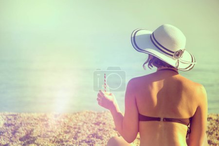 Foto de Mujer sentada en la arena de la playa, sosteniendo un zumo de naranja, llevando un bikini y un sombrero de paja y mirando hacia el horizonte - Imagen libre de derechos
