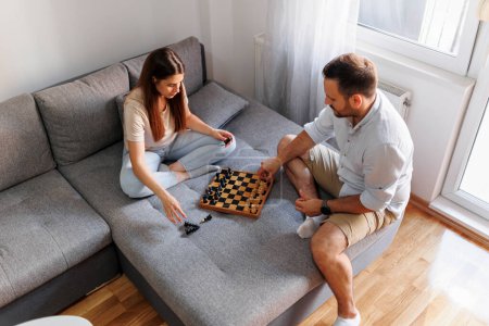 Téléchargez les photos : Vue grand angle du couple amoureux assis sur le canapé dans le salon, s'amuser à jouer aux échecs et profiter du temps libre à la maison - en image libre de droit
