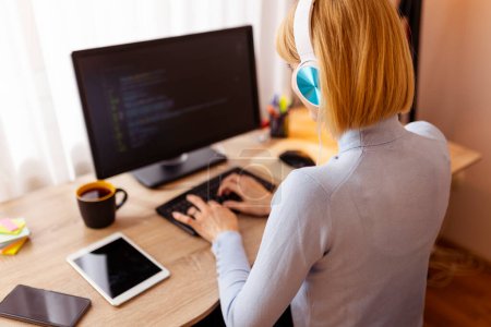 Photo for Female web developer sitting at her desk in home office wearing headset coding on desktop computer - Royalty Free Image