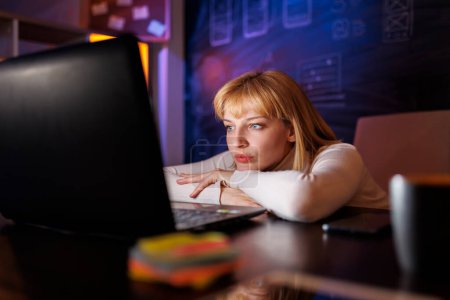 Photo for Beautiful young woman leaning at her desk in home office, looking at computer screen while working late at night - Royalty Free Image