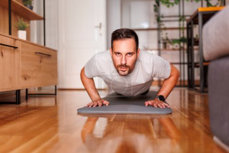 Téléchargez les photos : Beau homme actif travaillant à la maison, faisant des pompes sur tapis de yoga ; homme faisant de l'exercice à la maison le matin - en image libre de droit