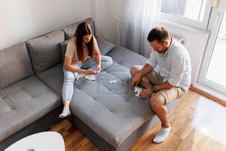 Photo for High angle view of couple in love sitting on couch in living room, having fun playing dominoes while spending their leisure time together at home - Royalty Free Image
