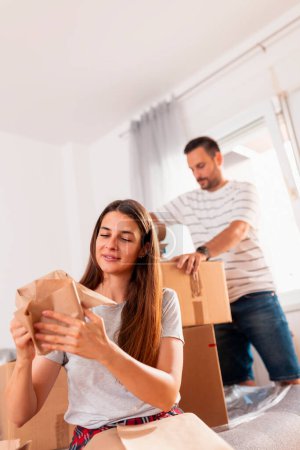 Photo for Couple in love packing things into cardboard boxes, getting ready for relocation - man taping boxes using packing machine while woman is wrapping fragile things into paper - Royalty Free Image
