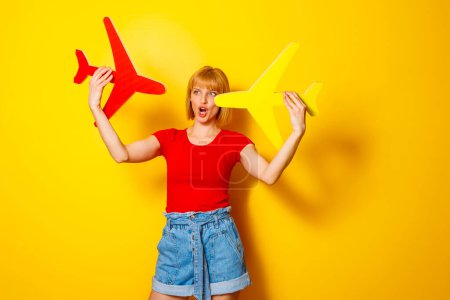 Photo for Woman in trendy summer clothes holding two cardboard planes on yellow color background, wodering about next travel destination; summer vacation and air travel concept - Royalty Free Image