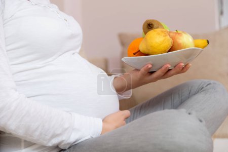 Foto de Acercamiento del vientre de una mujer embarazada mientras está sentada en un sofá en una sala de estar, y suavemente sosteniendo su vientre con una mano y un tazón de fruta en el otro - Imagen libre de derechos