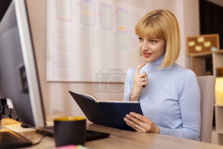 Téléchargez les photos : Femme assise à son bureau à la maison, écrivant dans le planificateur tout en prenant un cours éducatif en ligne - en image libre de droit