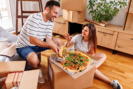 Téléchargez les photos : Couple amoureux emménager ensemble, prendre une pause de déballer des boîtes en carton, s'amuser tout en mangeant de la pizza pour le déjeuner - en image libre de droit