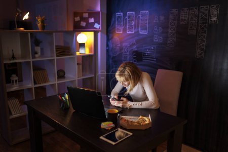 Photo for Woman sitting at her desk in home office using smart phone while working late at night - Royalty Free Image