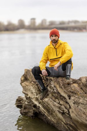 Téléchargez les photos : Homme assis sur un tronc d'arbre tombé, faisant une pause en faisant du jogging au bord de la rivière par un jour couvert d'automne - en image libre de droit