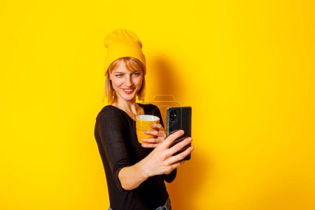 Photo for Portrait of beautiful blond woman wearing hat taking a selfie while drinking coffee on yellow color background with copy space - Royalty Free Image