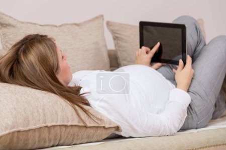 Téléchargez les photos : Jeune femme enceinte, relaxante allongée sur un canapé et surfant sur le net sur une tablette - en image libre de droit