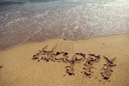 Téléchargez les photos : Mot heureux écrit à la main dans le sable sur la plage à côté de la ligne de flottaison - en image libre de droit
