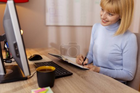 Téléchargez les photos : Femme assise à son bureau, prenant des notes dans le planificateur tout en ayant une conférence téléphonique liée au travail - en image libre de droit