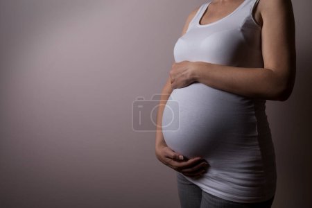 Téléchargez les photos : Gros plan d'une femme enceinte, isolée sur fond gris, tenant son ventre en douceur et protectrice - en image libre de droit