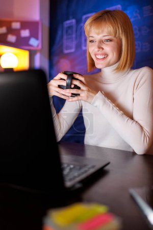 Photo for Woman sitting at her desk in home office, drinking coffee and using laptop computer while working late - Royalty Free Image