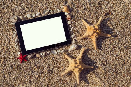 Foto de Marco de fotos hecho de conchas marinas y guijarros con dos estrellas de mar colocadas junto a él y tableta de computadora dentro del marco con pantalla en blanco en la playa - Imagen libre de derechos