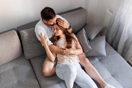 Photo for High angle view of a beautiful young couple in love relaxing at home, lying cuddling and hugging on the couch, men kissing woman in forehead both with eyes closed - Royalty Free Image