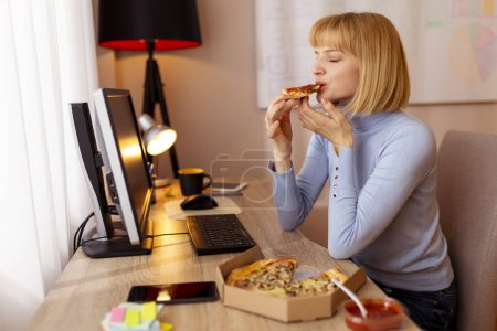 Photo for Woman sitting at her desk an office, taking a lunch break eating pizza - Royalty Free Image