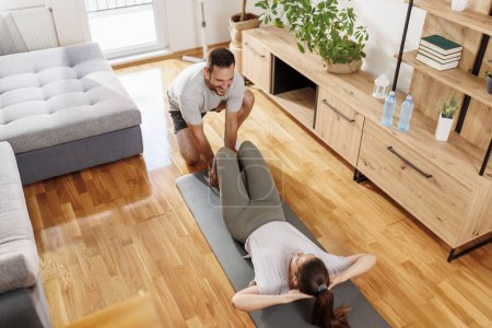 Foto de Pareja enamorada haciendo ejercicio juntos en casa, mujer acostada en una esterilla de yoga haciendo crujidos mientras el hombre sostiene sus piernas y cuenta - Imagen libre de derechos