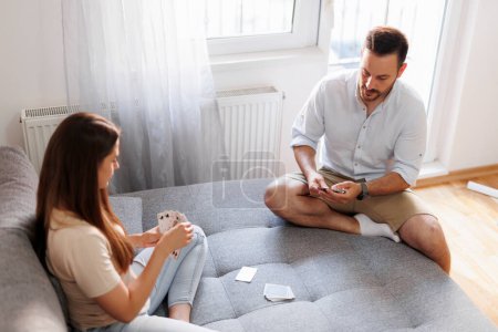 Téléchargez les photos : Beau jeune couple amoureux profiter de leur temps libre à la maison ensemble et avoir du plaisir à jouer à des jeux de cartes - en image libre de droit