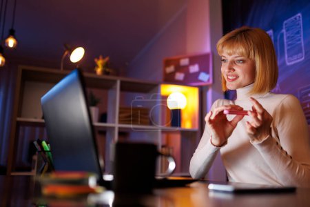 Téléchargez les photos : Belle jeune femme prenant une pause tout en travaillant tard au bureau à la maison, assis à son bureau, en utilisant une lime à ongles et regarder un film en ligne - en image libre de droit