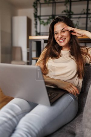 Foto de Hermosa mujer joven acostada en el sofá con el ordenador portátil en su regazo, riendo mientras tiene videollamada con amigos - Imagen libre de derechos