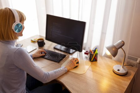 Photo for Female software developer sitting at her desk in home office wearing headset coding on desktop computer while working remotely from home - Royalty Free Image