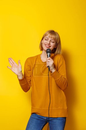 Téléchargez les photos : Portrait de jeune femme joyeuse s'amusant tenant micro et chantant karaoké isolé sur fond de couleur jaune - en image libre de droit